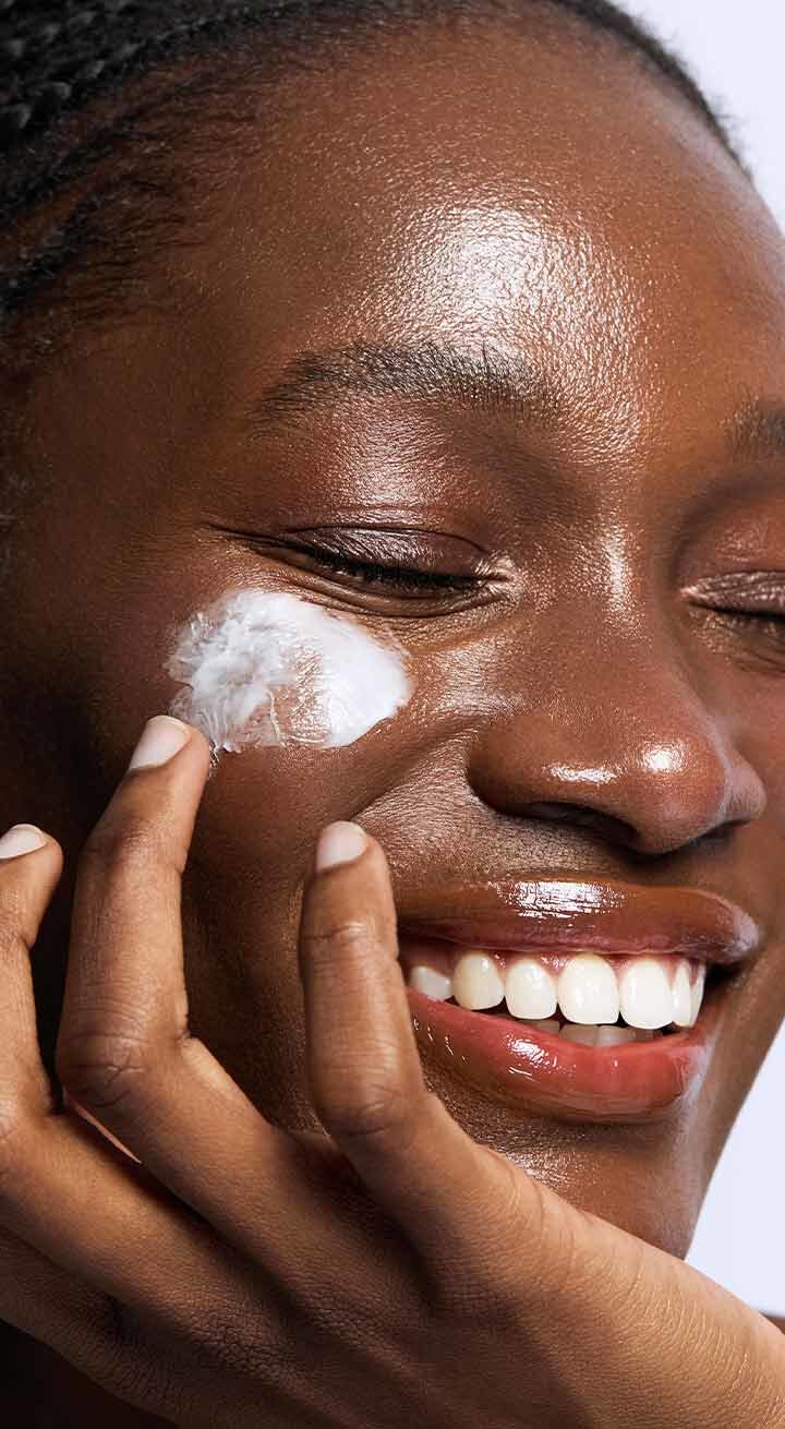 A smiling person with dark skin gently applies a generous dollop of Laneige's Cica Sleeping Mask to their cheek, showcasing the mask's creamy texture and highlighting its moisturizing benefits.