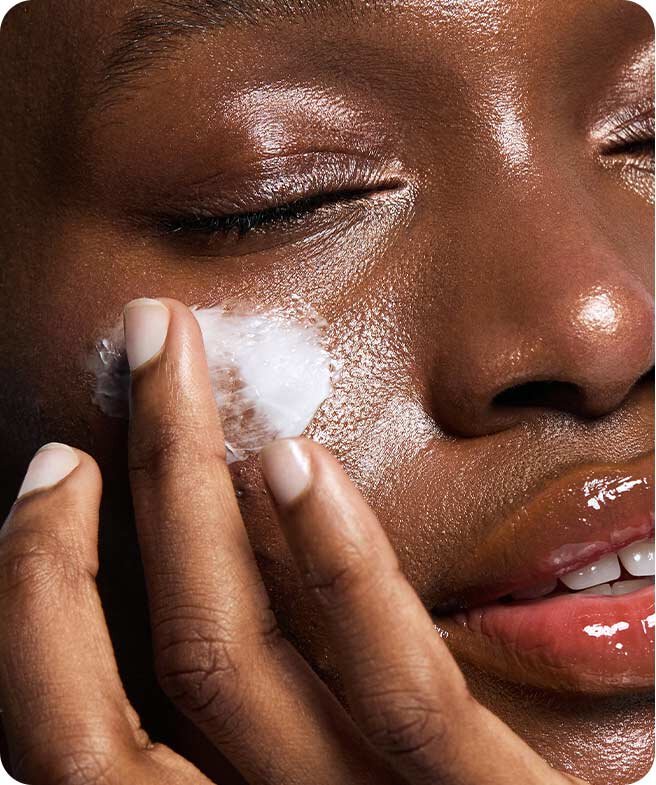 A person with dark skin gently applies a generous amount of Laneige's Cica Sleeping Mask to their cheek, eyes closed in a relaxed expression, showcasing the mask's creamy texture and the product's soothing effect on the skin.
