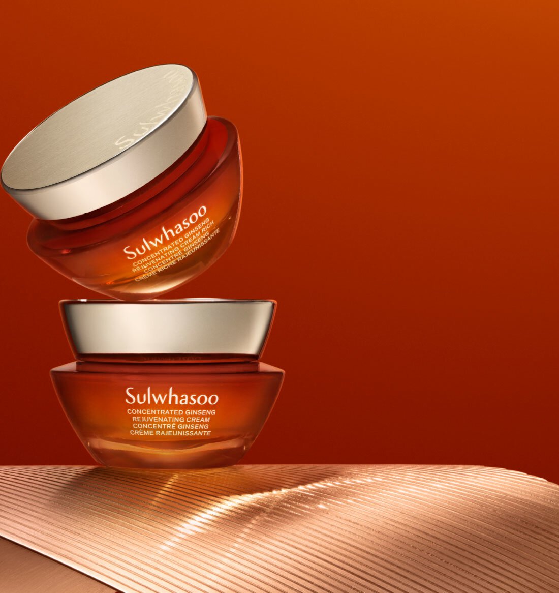 Two jars of Sulwhasoo Concentrated Ginseng Rejuvenating Cream Rich, one slightly levitated above the other, showcase the product's luxurious texture and rich color against a warm orange backdrop.